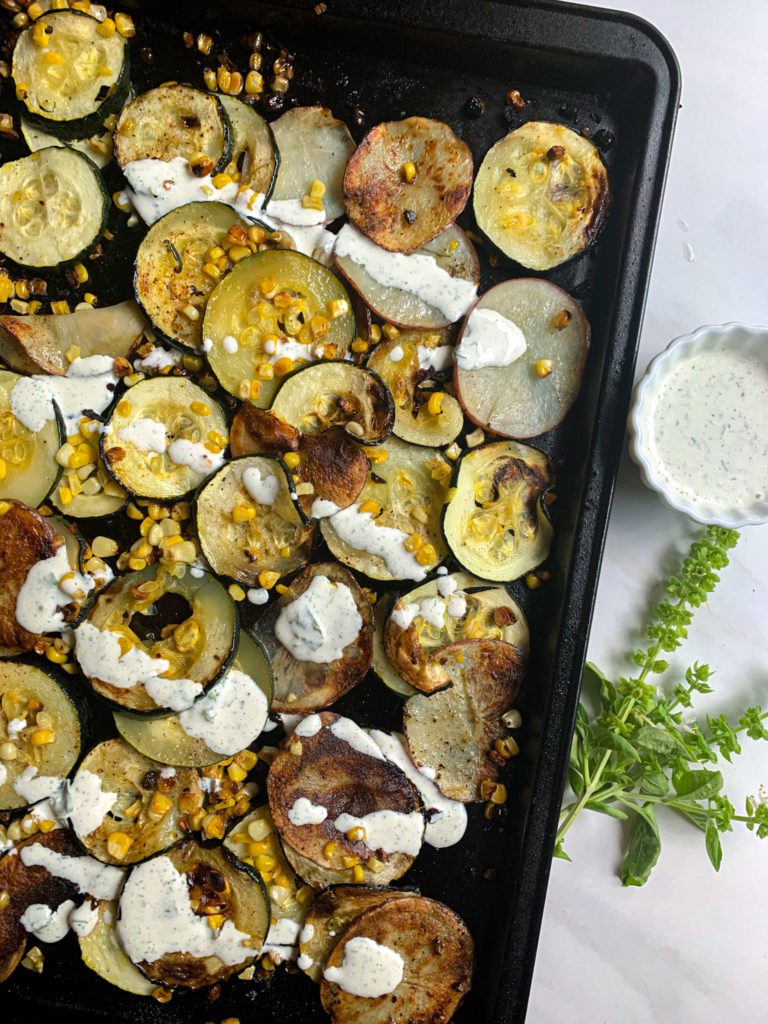 Sheet pan with thinly sliced and layered garden fresh zucchini and potato topped with sweet corn cut off the cob and drizzled with homemade basil ranch.