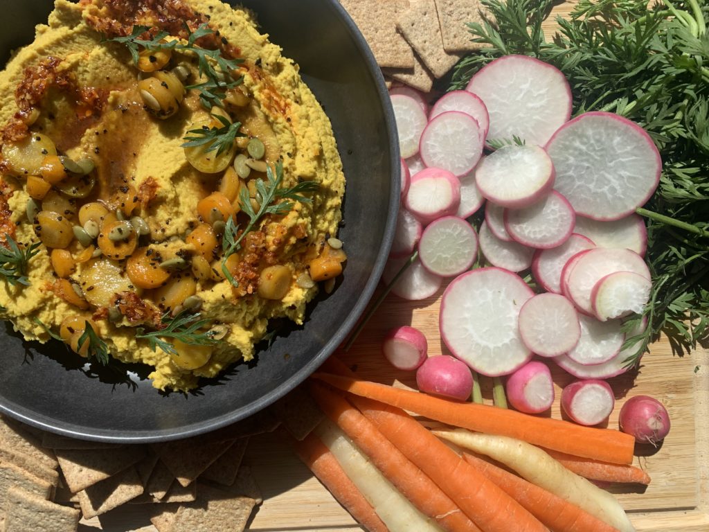 A bowl of yellow carrot hummus with carrot toppings, chili oil, mike's hot honey, toasted pepitas, and roasted carrots on top. Served with crackers, carrots, and farmer's market fresh radishes. Alongside this pictures is a recipe for Farmers Market Carrot Hummus.