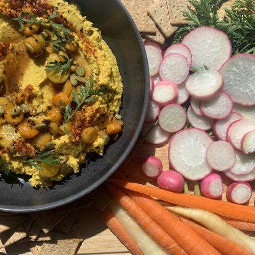 A bowl of yellow carrot hummus with carrot toppings, chili oil, mike's hot honey, toasted pepitas, and roasted carrots on top. Served with crackers, carrots, and farmer's market fresh radishes. Alongside this pictures is a recipe for Farmers Market Carrot Hummus.