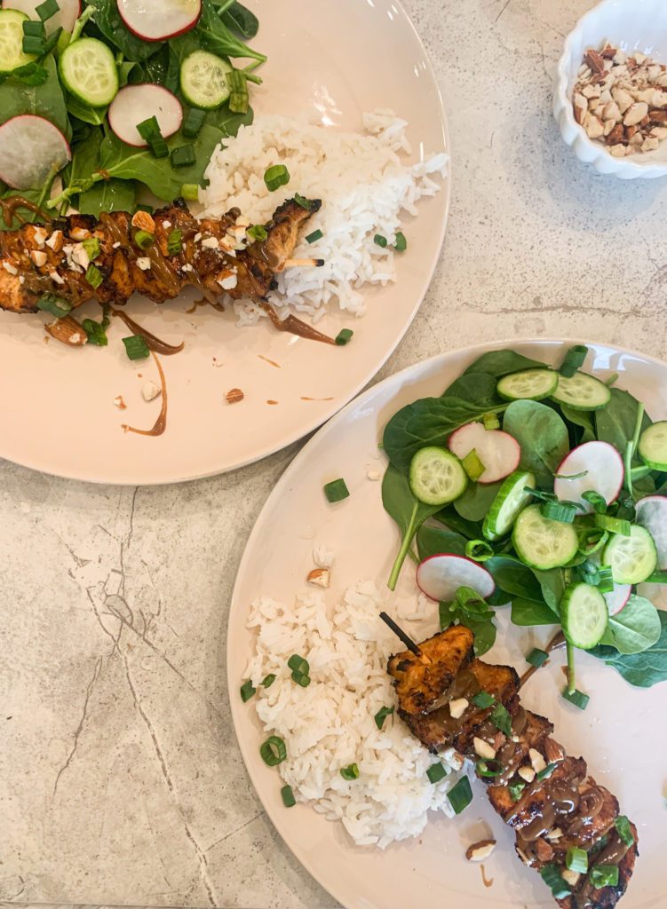 Spicy Asian glazed chicken on wooden skewers topped with creamy peanut sauce and sliced almonds. On the side is a mound of white rice and salad with cucumbers, radishes, and spinach.