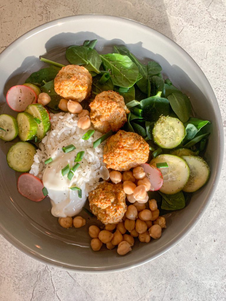 A ceramic bowl with spinach, garbanzo beans, white rice, pickled cucumber and radish salad, topped with Gluten Free Greek Chicken Meatballs and Greek yogurt. Under the picture is a recipe for the Gluten Free Greek Chicken Meatballs.