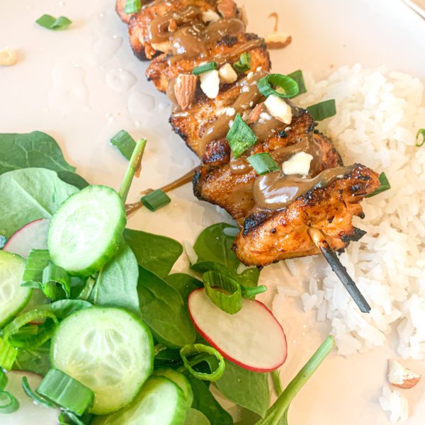 Spicy Asian glazed chicken on wooden skewers topped with creamy peanut sauce and sliced almonds. On the side is a mound of white rice and salad with cucumbers, radishes, and spinach.