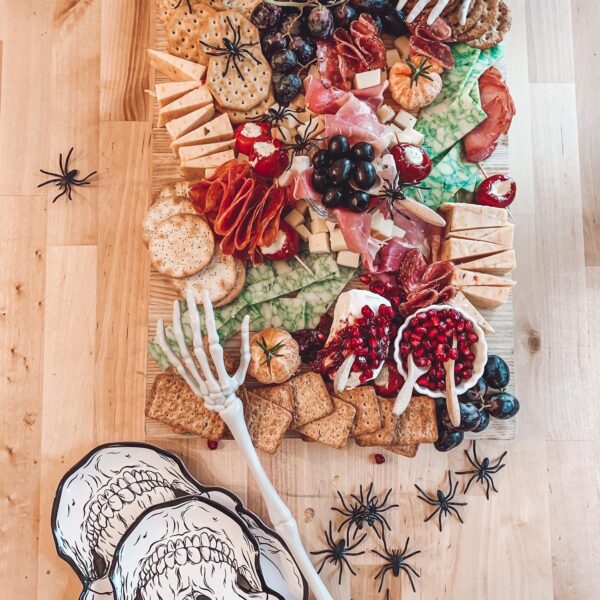 Pictures of an extravagant Halloween inspired charcuterie board. The board is overflowing with meats, cheeses, crackers and spiders! There are plastic spiders and skeleton hands added to the board for a spooky touch.