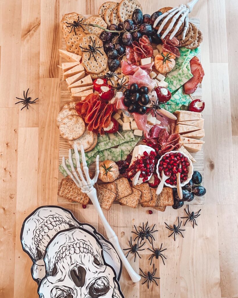 Pictures of an extravagant Halloween inspired charcuterie board. The board is overflowing with meats, cheeses, crackers and spiders! There are plastic spiders and skeleton hands added to the board for a spooky touch.