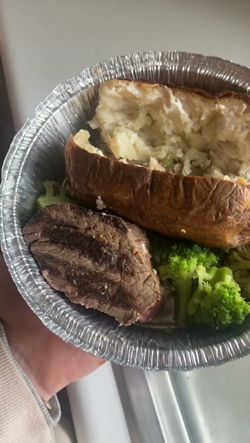 Half of a leftover filet with steamed broccoli and a baked potatoes in a leftover container from a restaurant.
