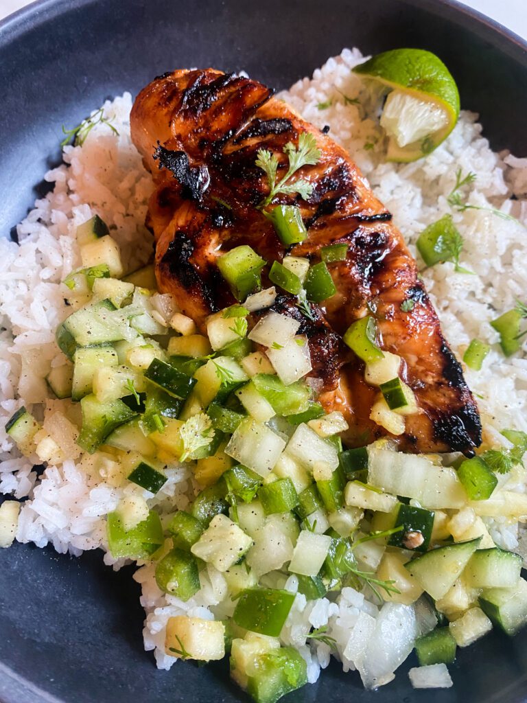Bowl with white rice topped with grilled pineapple marinated chicken and pineapple salsa with cucumbers, pineapple, green bell peppers, onion, and cilantro.  Squeezed lime wedge on the side. 