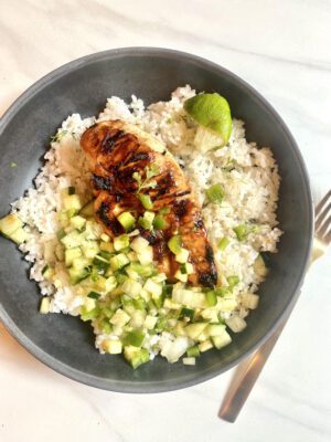 Bowl with white rice topped with grilled pineapple marinated chicken and pineapple salsa with cucumbers, pineapple, green bell peppers, onion, and cilantro. Squeezed lime wedge on the side.