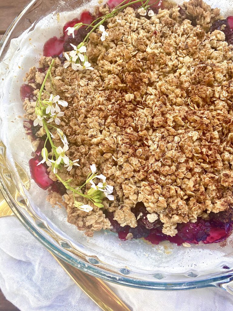 Pie dish filled with blackberry filling and an oat and flax seed crumble on top. Laid on a rustic table setting with flowers on top of the baked blackberry and honey crumble.