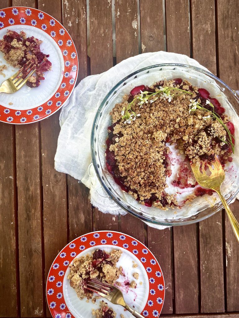 Pie dish filled with blackberry filling and an oat and flax seed crumble on top. Laid on a rustic table setting with flowers on top of the baked blackberry and honey crumble. Two vintage tea plates with small servings of the crumble and forks that have already taken a bite are set next to the dish.