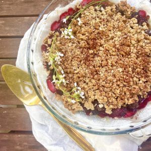 Pie dish filled with blackberry filling and an oat and flax seed crumble on top. Laid on a rustic table setting with flowers on top of the baked blackberry and honey crumble