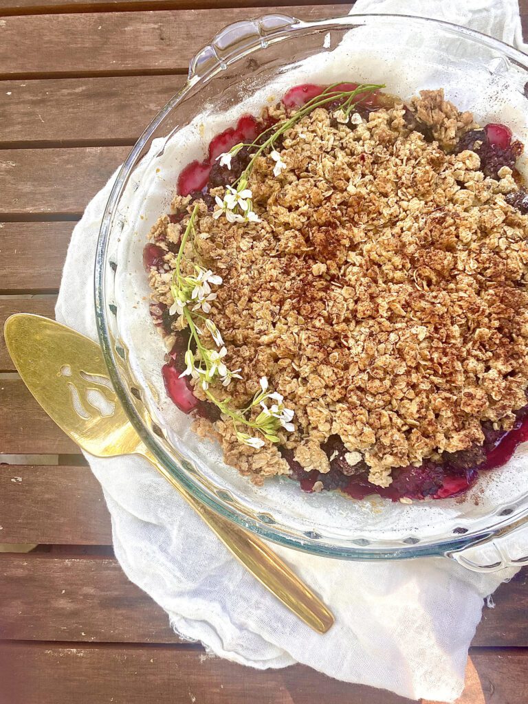 Pie dish filled with blackberry filling and an oat and flax seed crumble on top. Laid on a rustic table setting with flowers on top of the baked blackberry and honey crumble