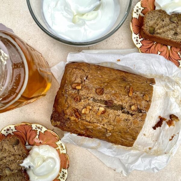 Fresh baked Burnt Butter & Brown Sugar Banana Bread in the middle of the photo with two pieces sliced off and placed on two floral vintage plates with a dollop of Greek yogurt served alongside the banana bread and drizzled with local honey right from a mason jar.