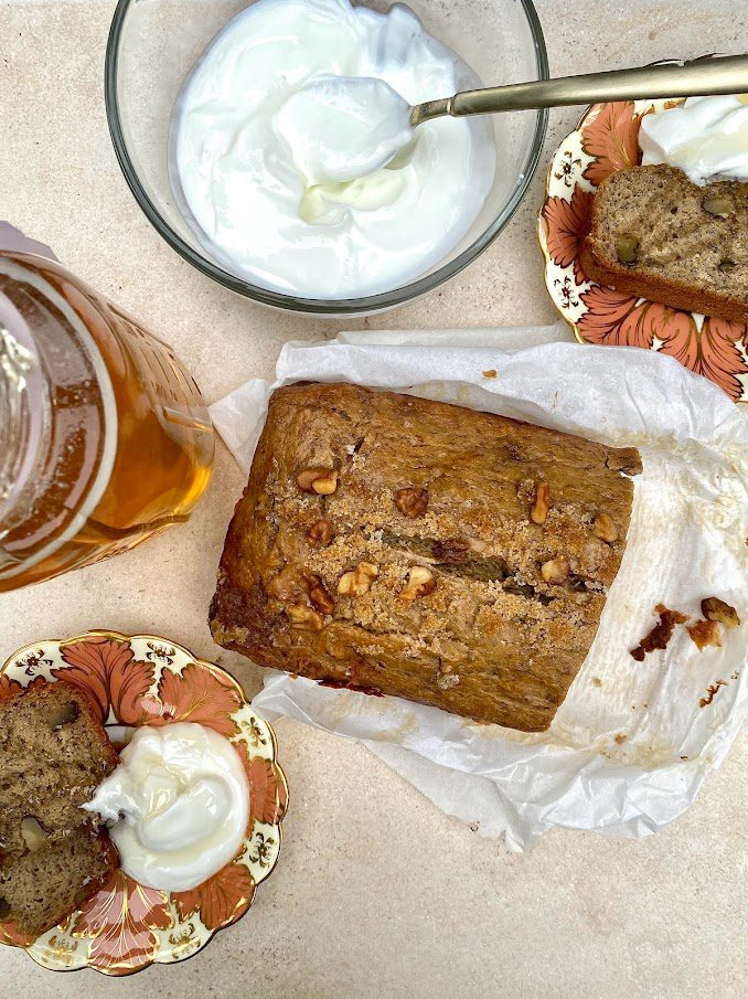 Fresh baked Burnt Butter & Brown Sugar Banana Bread in the middle of the photo with two pieces sliced off and placed on two floral vintage plates with a dollop of Greek yogurt served alongside the banana bread and drizzled with local honey right from a mason jar.